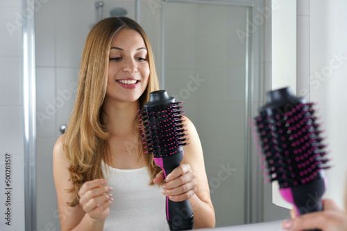 Smiling girl holds round brush hair dryer in her bathroom at home. Young woman looking satisfied her salon one-step brush hair dryer and volumizer. photo