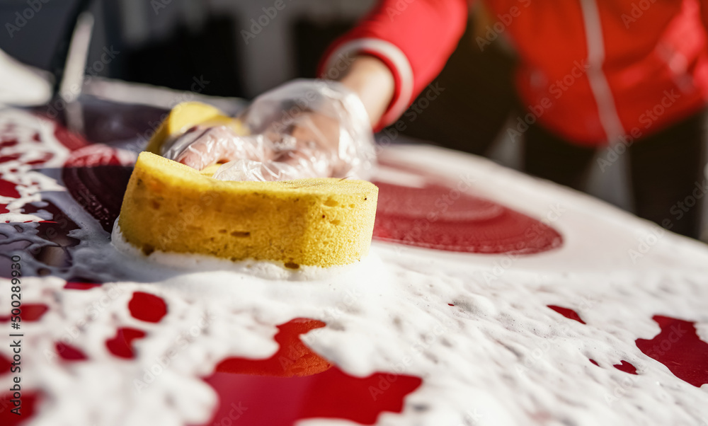 Young woman cleaning front hood of her car, closeup detail on yellow sponge held by hand in plastic glove Young woman cleaning front hood of her car, closeup detail on yellow sponge held by hand in plastic glove