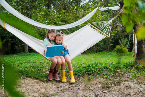 Outdoor activities with kids. Girls siting in hammock and reading a book in the garden. Friendship and childhood. Summertime concept.
