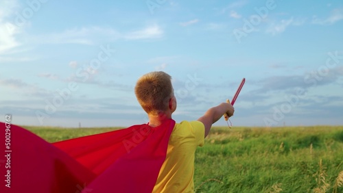Child boy plays superhero. Child Game, Imagination. Boy, child in red cloak go with sword raised in his hand up sword, on field, depicting medieval knight. Child waves toy sword, childhood dreams. © zoteva87
