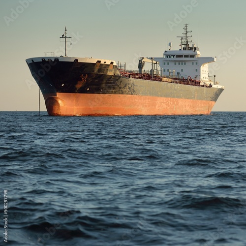 Large cargo ship (chemical tanker, 184 meters length) sailing in an open sea at sunset. Golden sunlight. Freight transportation, fuel, power generation, nautical vessel, logistics, economy, industry