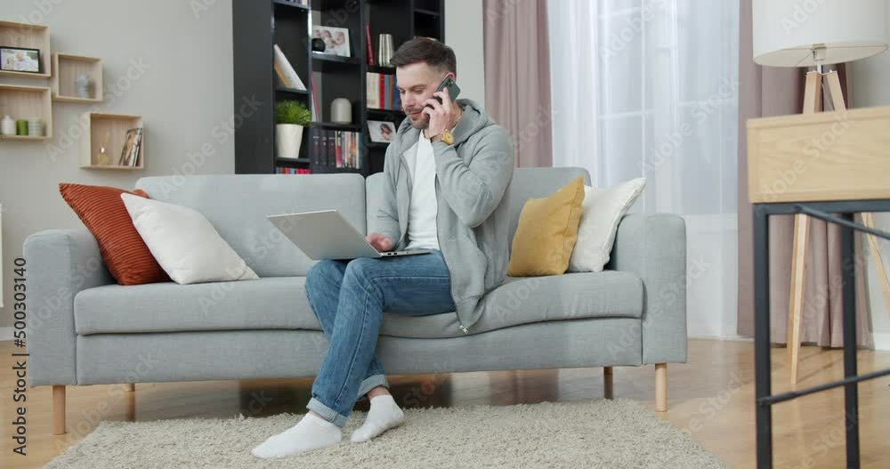 Handsome caucasian man talking on mobile phone and typing on wireless laptop while sitting on comfy couch. Modern gadget for domestic lifestyles. Handsome caucasian man talking on mobile phone and typing on wireless laptop while sitting on comfy couch. Modern gadget for domestic lifestyles.