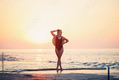 Attractive young girl with long hair poses in front of the camera on the beach. She is wearing a black swimsuit. Golden sunset light