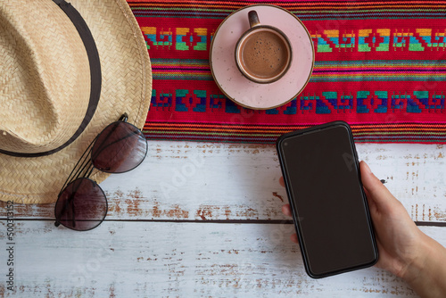 Celular, taza de café sombrero y lentes con mantel artesanal mexicano