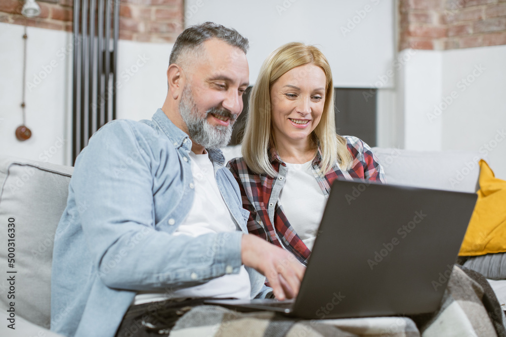 Obraz premium Smiling married couple of mature man and woman spending leisure time at home with modern laptop. Caucasian family sitting on couch and watching some movie.