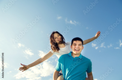 A young man carries his beautiful smiling woman on his back in the park. Couple in love having fun together.