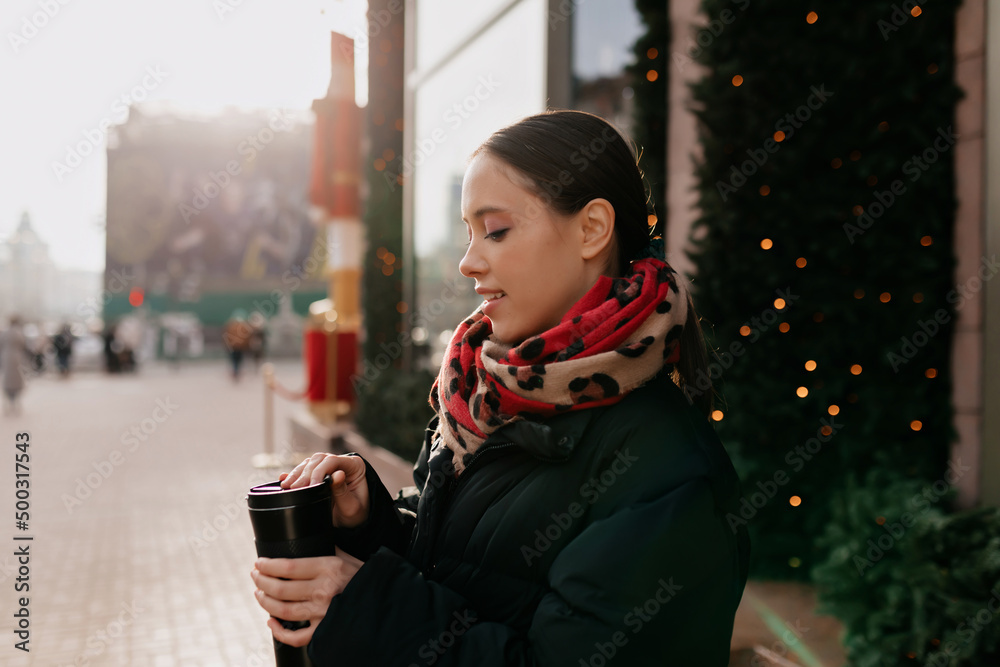 Profile portrait of brunette girl wearing warm dark jacket and scarf is enjoying coffee and walking outdoor in sunlight and warm winter day