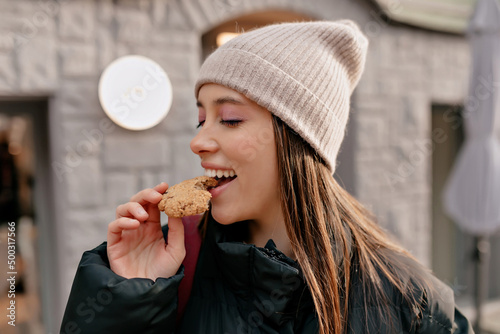 Happy excited girl with dark hair wearing beige cap and dark jacket is bitting cookies on the outdoor cafe in sunshine
