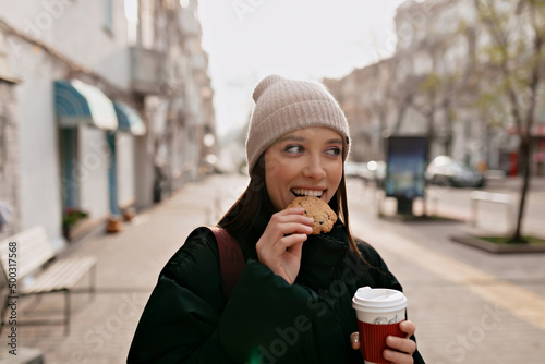 Portrait of lovely cute girl with dark hair is bitting cookies and holding coffee while walking around the city in sunny day