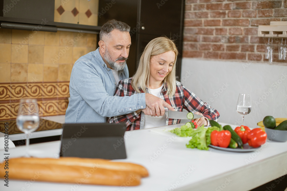 Happy middle aged family in casual attire using digital tablet and cooking healthy fresh meal on kitchen. Caucasian preparing meal from fresh organic vegetables. Happy middle aged family in casual attire using digital tablet and cooking healthy fresh meal on kitchen. Caucasian preparing meal from fresh organic vegetables.