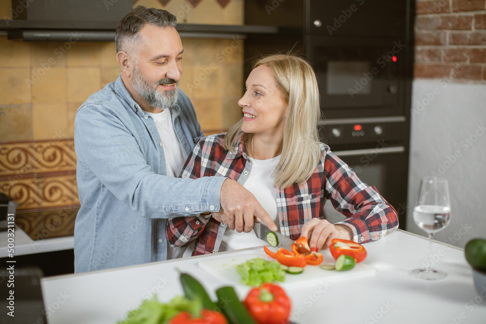 Happy mature couple in love chopping fresh vegetables on kitchen and using digital tablet. Caucasian family in casual wear enjoying common time at cozy home.