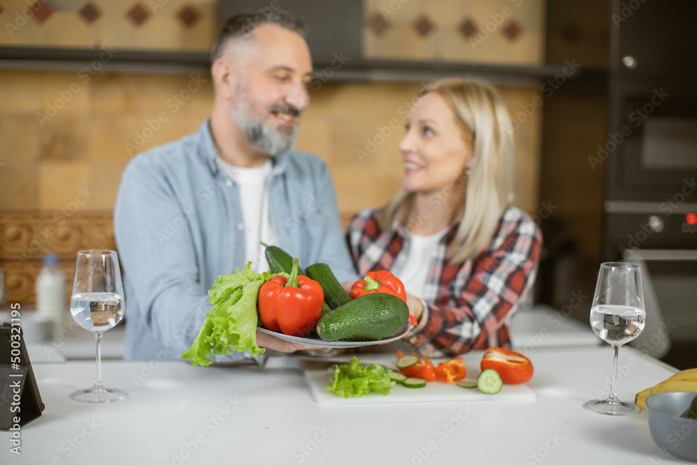 Mature married couple in casual clothes holding plate with healthy eco vegetables while standing on kitchen and smiling to each other. Concept of family and lifestyles. Mature married couple in casual clothes holding plate with healthy eco vegetables while standing on kitchen and smiling to each other. Concept of family and lifestyles.
