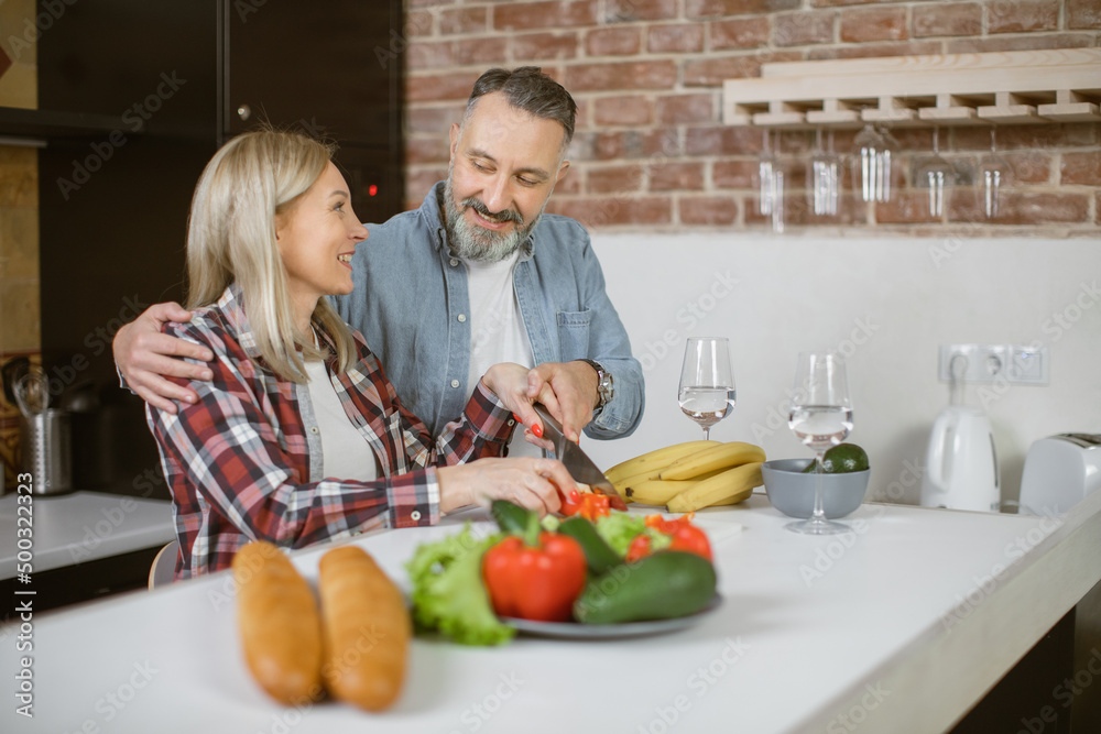 Handsome mature man and charming woman enjoying cooking process at modern domestic kitchen. Happy family using fresh organic vegetables for healthy meal.