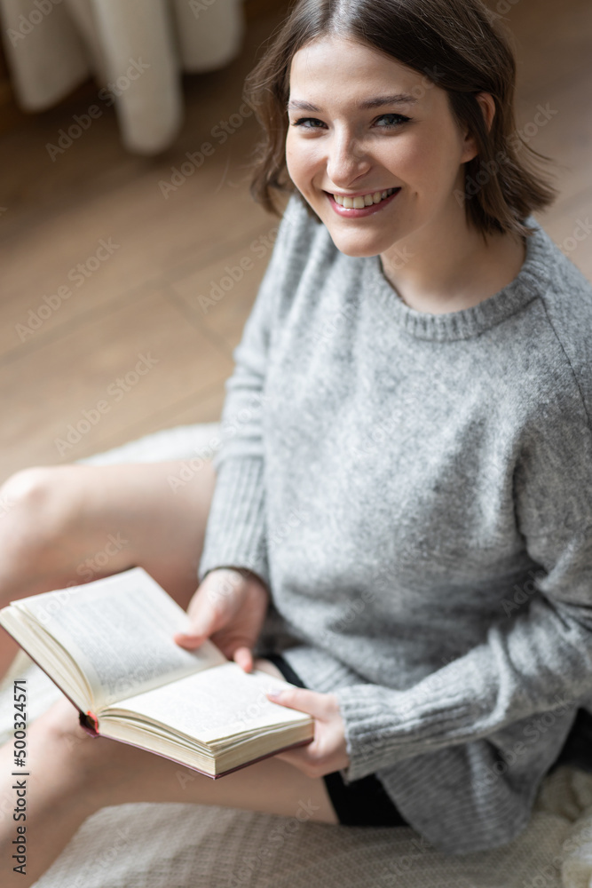 Beautiful young woman relaxed on the floor in the living room at home reading a book. Daylight. Female in a grey sweater filling comfortable at her house. Weekend Beautiful young woman relaxed on the floor in the living room at home reading a book. Daylight. Female in a grey sweater filling comfortable at her house. Weekend