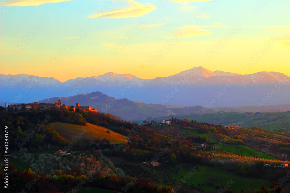 Scenic landscape view from Monte Vidon Corrado at a small town illuminated by beams of the setting sun and surrounded by green hills and the Apennines with snow-capped peaks