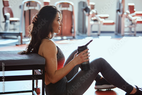 Принти на полотні A sportswoman sits on the floor in a gym and drinks a protein shake for strength