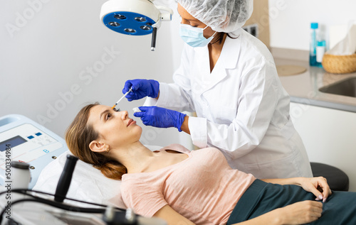 Professional female beautician in mask performing face contouring injections to european woman patient at cosmetology clinic