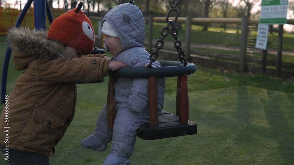 Obraz premium Funny Little Preschool Chubby Girl Older Sister Rides Younger Newborn Infant Brother Boy On Swing. Baby Playing On Playground. Kids Entertainment, Childhood, Child Development, Happy Family Concept