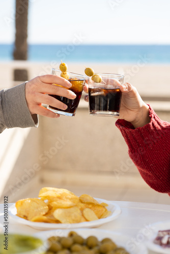 Close up of two hands toasting with their glasses of vermouth on a table full of snacks in front of a beach on a sunny day. Two people with no faces, toasting with some drinks and aperitivos outdoors.