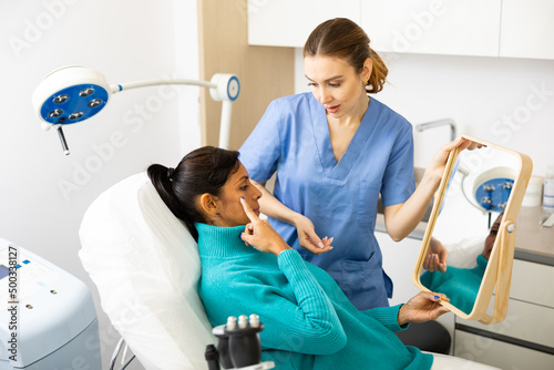 Hispanic woman holding mirror and pointing finger on her face during cosmetologist s examination in clinic.
