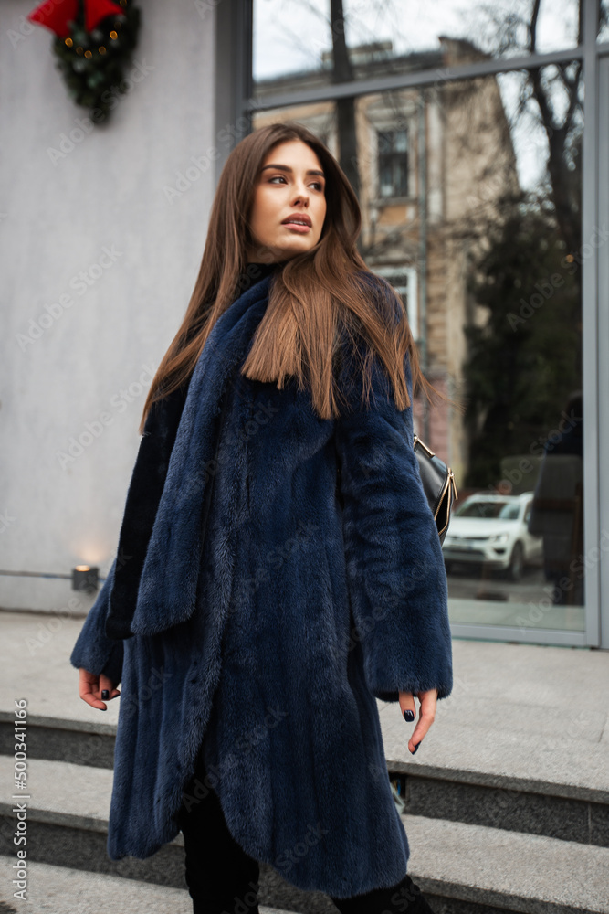 Girl posing on road on winter background. Glamorous funny young woman with smile wearing stylish blue long fur coat . Fur and fashion concept.