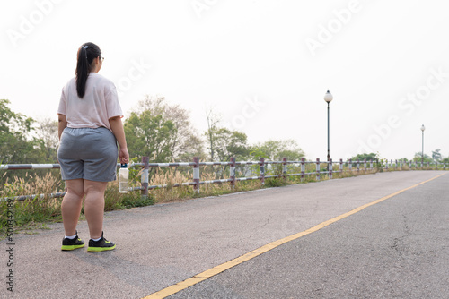 Rear view angle  wide shot of unrecognizable fat woman standing  holding sports water bottle  wearing sportswear  looking away at long country road. Goal setting  weight loss  self resolution concept.