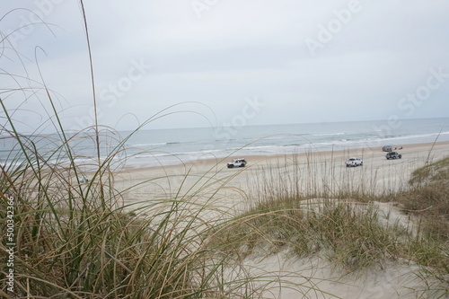 Fototapeta Naklejka Na Ścianę i Meble -  Parked Cars, People, on Sandy Beach, Ocean Waves, Blue Sky and Dunes