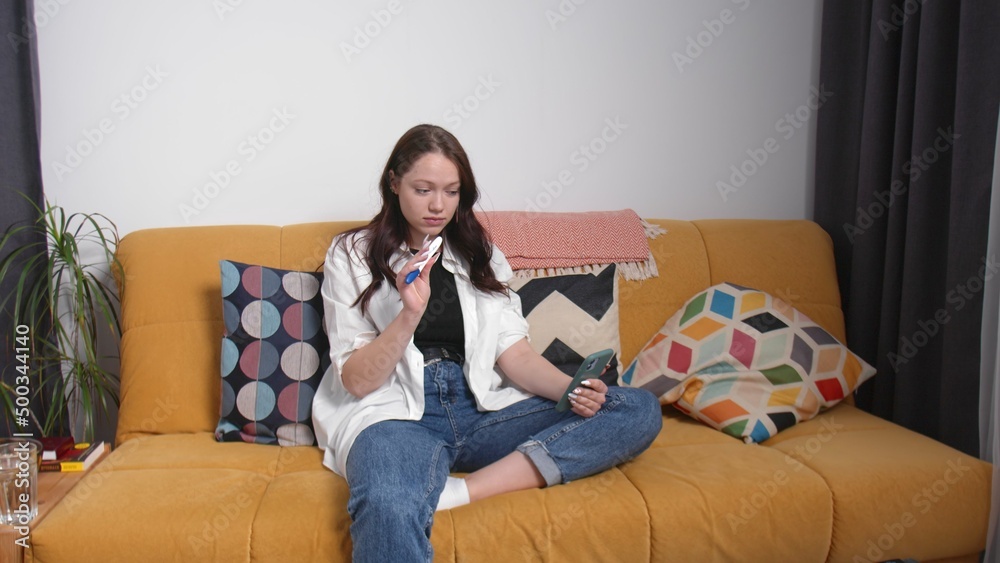 Obraz premium A shocked young woman is sitting on the couch and talking via video link on her phone and showing a pregnancy test, a young woman is pregnant. Early pregnancy