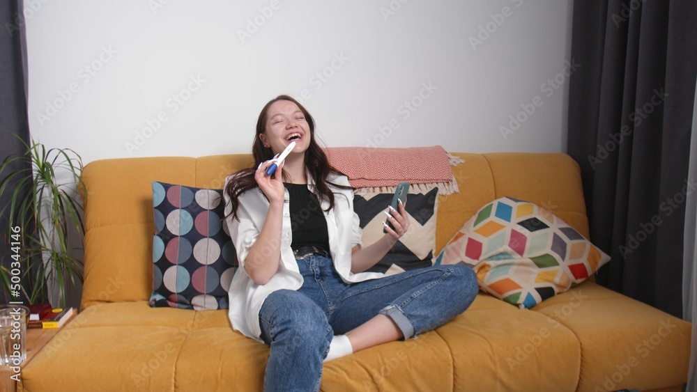 Obraz premium A happy young woman is sitting on the couch and talking via video link from her phone and showing a pregnancy test. A young woman is pregnant. Early pregnancy