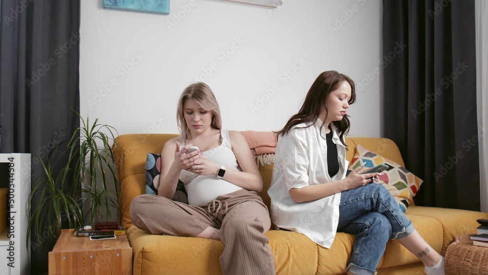 Young lesbians are offended at each other, sitting on the couch and looking at their phones. Pregnant lesbian woman. LGBT. An unconventional family. Young lesbians are offended at each other, sitting on the couch and looking at their phones. Pregnant lesbian woman. LGBT. An unconventional family.