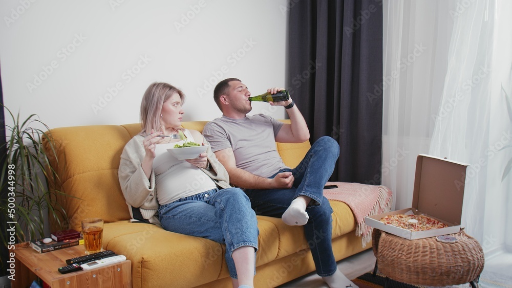 A pregnant young woman eats healthy food, and a man drinks beer from a bottle. A young married couple is relaxing on the couch and watching TV. Young family A pregnant young woman eats healthy food, and a man drinks beer from a bottle. A young married couple is relaxing on the couch and watching TV. Young family