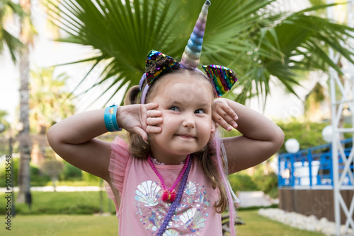 Portrait of a 4 year old girl dressed in a rainbow dress and a unicorn headband against the backdrop of palm trees