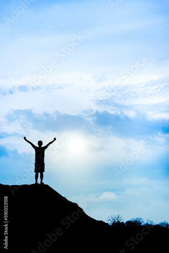 Silhouette of climber success at the top of mountain with cloudy blue sky. A man standing and raising arms in the air with bright morning sun. Achieving a goal, motivation and positive energy concept.