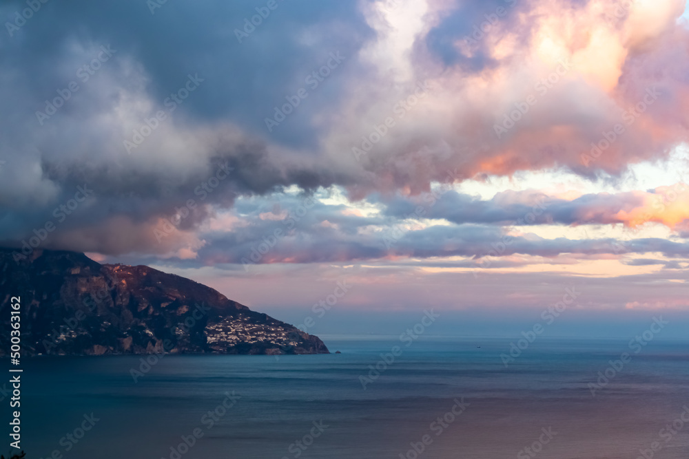 Distant view on the coastal town Positano in Campania, Italy, Europe. Sunset over Mediterranean sea on Amalfi Coast. Golden hour sunlight is touching the building of the fishermen village. Red clouds Distant view on the coastal town Positano in Campania, Italy, Europe. Sunset over Mediterranean sea on Amalfi Coast. Golden hour sunlight is touching the building of the fishermen village. Red clouds
