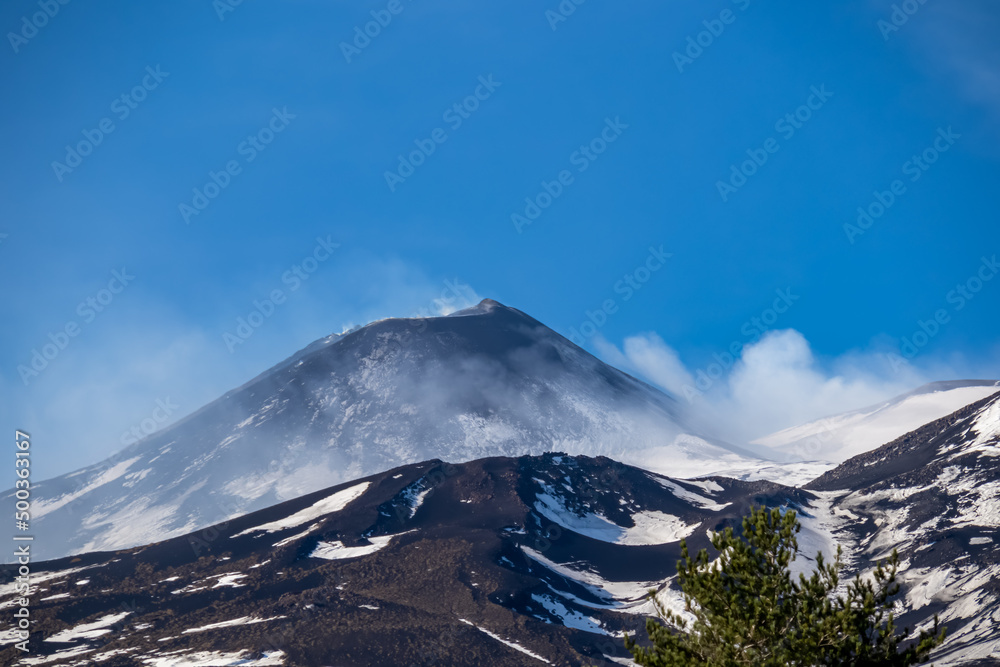 Panoramic view on north eastern flank of volcano mount Etna, in Sicily, Italy, Europe. Summit is covered with snow. Landscape is black brown volcanic sand, bare terrain. Smoke is around the craters