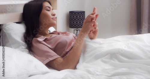 Brunette woman dressed in pink silk pajamas, lying on a white bed in the bedroom, smiles and laughs while reading a message on her cell phone. Asian woman rejoices at the good news