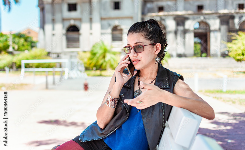 Fototapeta premium Urban style girl sitting on a bench calling on the phone, An attractive girl sitting talking on the cell phone, Girl sitting in a park calling on the phone