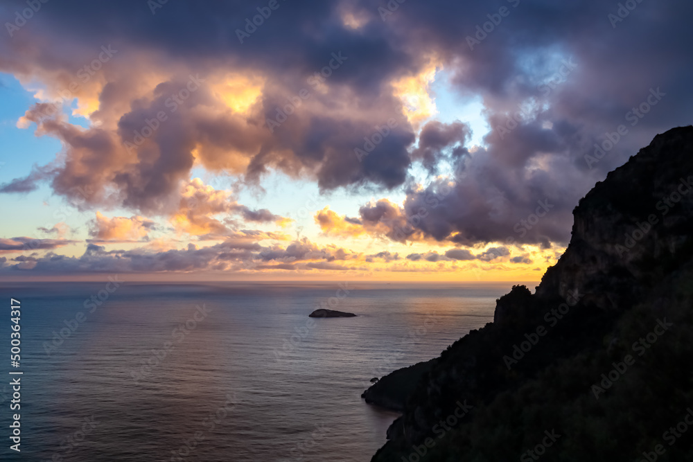 Sunset over the island of Scoglio Vetara on the Mediterranean sea at Amalfi Coast, Campania, Italy, Europe. Golden hour sunlight on surface of Tyrrhenian Sea. Red orange clouds emerging. Coastline
