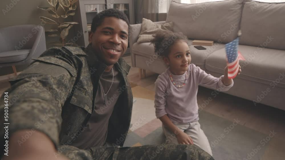 Handheld POV slowmo of young African American army officer in military uniform and his pretty little daughter with US flag in hands smiling at camera while taking selfie portrait at home