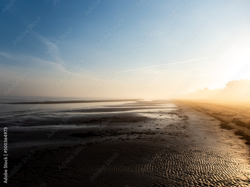 Beautiful landscape of peaceful Baltic sea beach at the sunrise in early foggy and misty morning with warm golden hour light from. Wet sand reflecting light with white mist in the air