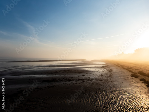 Beautiful landscape of peaceful Baltic sea beach at the sunrise in early foggy and misty morning with warm golden hour light from. Wet sand reflecting light with white mist in the air © KristineRada