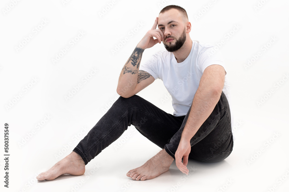 Young bearded man with short dark hair in white T-shirt, black jeans, sitting on white floor, leaning head on hand. Young bearded man with short dark hair in white T-shirt, black jeans, sitting on white floor, leaning head on hand.