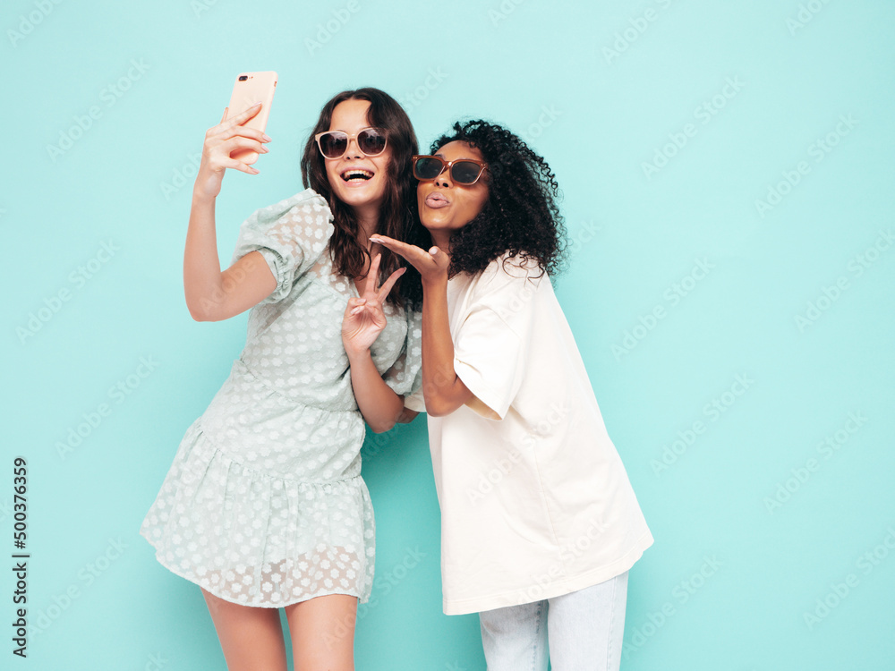Two young beautiful smiling international hipster female in trendy summer clothes. Sexy carefree women posing near blue wall in studio. Positive models having fun in sunglasses. Taking selfie photos Two young beautiful smiling international hipster female in trendy summer clothes. Sexy carefree women posing near blue wall in studio. Positive models having fun in sunglasses. Taking selfie photos