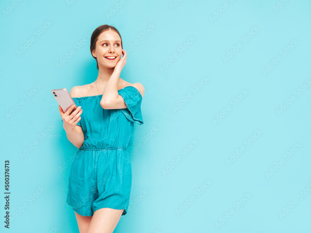 Young beautiful smiling female in trendy summer blue overalls. Sexy carefree woman posing near wall in studio. Positive brunette model looking at cellphone screen. Holding smartphone and using apps Young beautiful smiling female in trendy summer blue overalls. Sexy carefree woman posing near wall in studio. Positive brunette model looking at cellphone screen. Holding smartphone and using apps