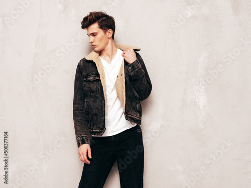 Portrait of handsome confident stylish hipster lambersexual model.Man dressed in black jacket and jeans. Fashion male posing in studio near grey wall