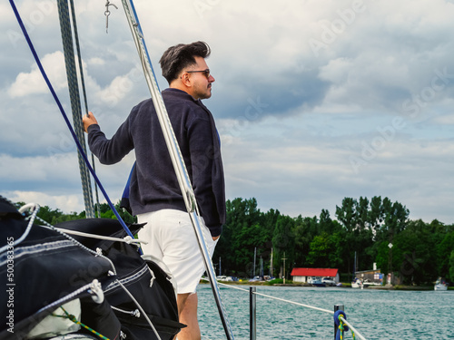 handsome person standing on a sailing yacht bow and looking a side in a cold day on a lake, back view