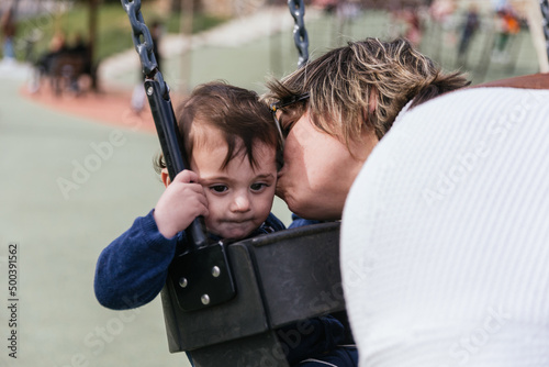 Lovely mom kissing her infant boy sitting on a swing at the playground