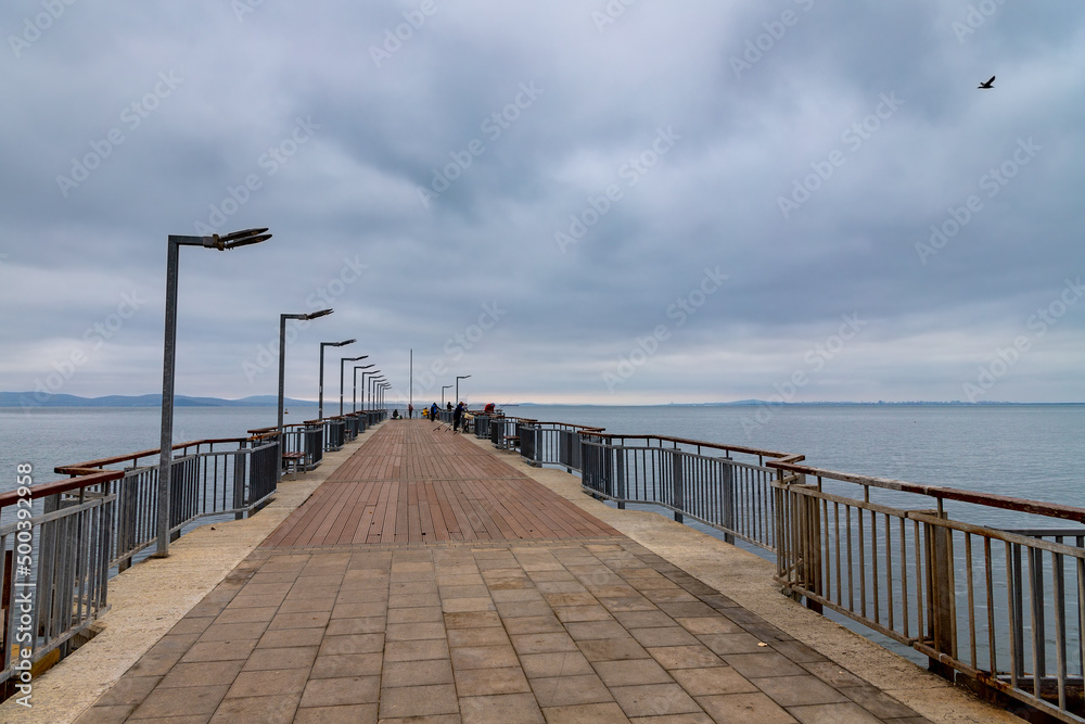 Perspective view of a wooden pedestrian bridge at sea, cloudy sky, and horizon in the background.