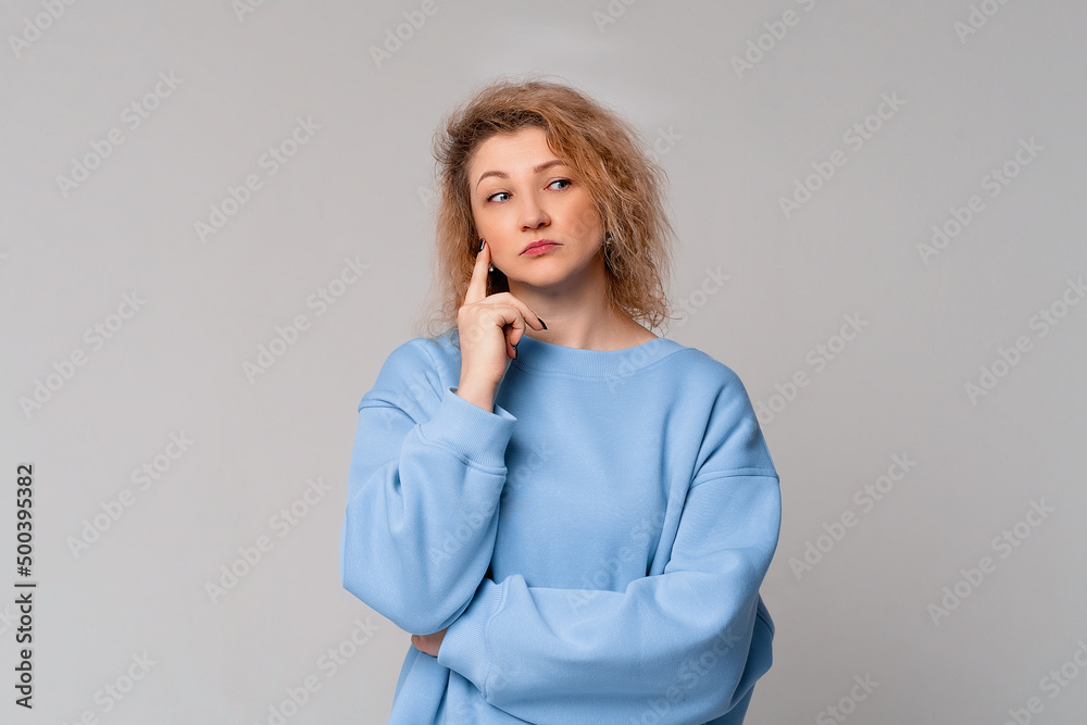 modern mature woman with curly blonde hair pointing to head with one finger, great idea or thought, good memory, standing in trendy blue sweatshirt over light grey background modern mature woman with curly blonde hair pointing to head with one finger, great idea or thought, good memory, standing in trendy blue sweatshirt over light grey background