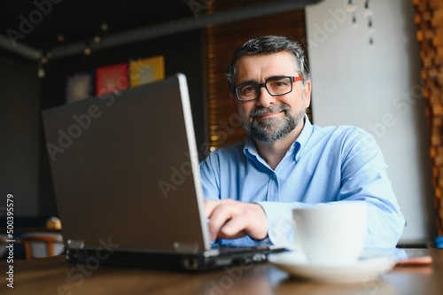 Mature businessman drinking coffee in cafe. Portrait of handsome man wearing stylish eyeglasses using laptop, looking at camera, smiling. Coffee break concept